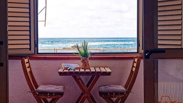 Una habitación con una mesa de madera y dos sillas, una planta en maceta y vistas al océano a través de contraventanas abiertas.