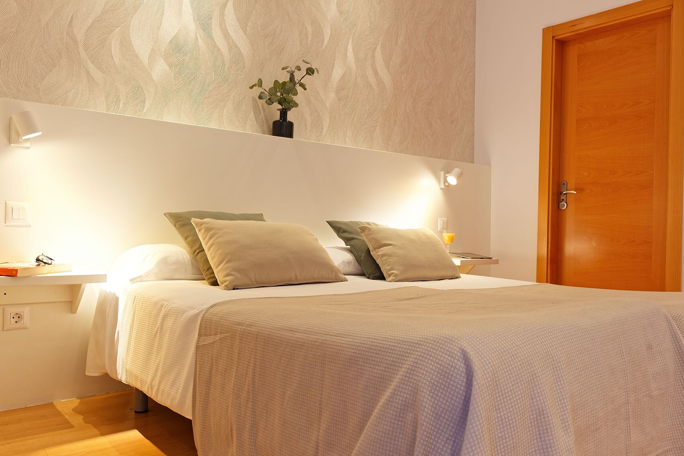 Una cama bien hecha con cubrecama blanco y beige, dos almohadas y una pequeña planta en maceta sobre una mesita de noche.