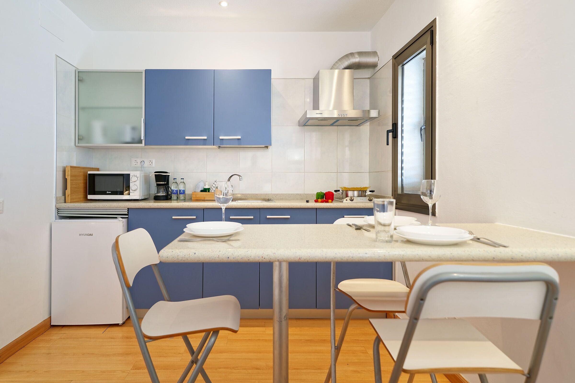 Cocina compacta con armarios azules, microondas, refrigerador pequeño y una mesa de comedor con sillas.