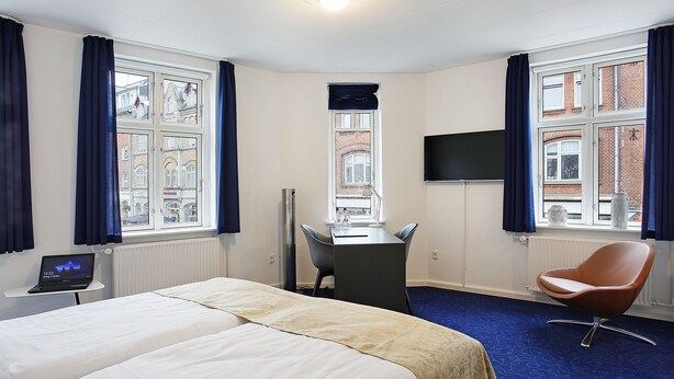 Ein Hotelzimmer mit Bett, Schreibtisch, Stuhl, Fernseher und einem Fenster mit Vorhängen.