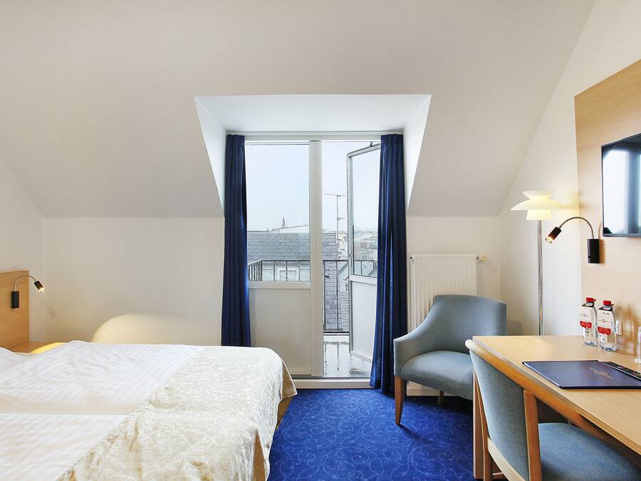 A hotel room with a bed, a desk, a chair, a TV, and a view of the city through a large window.