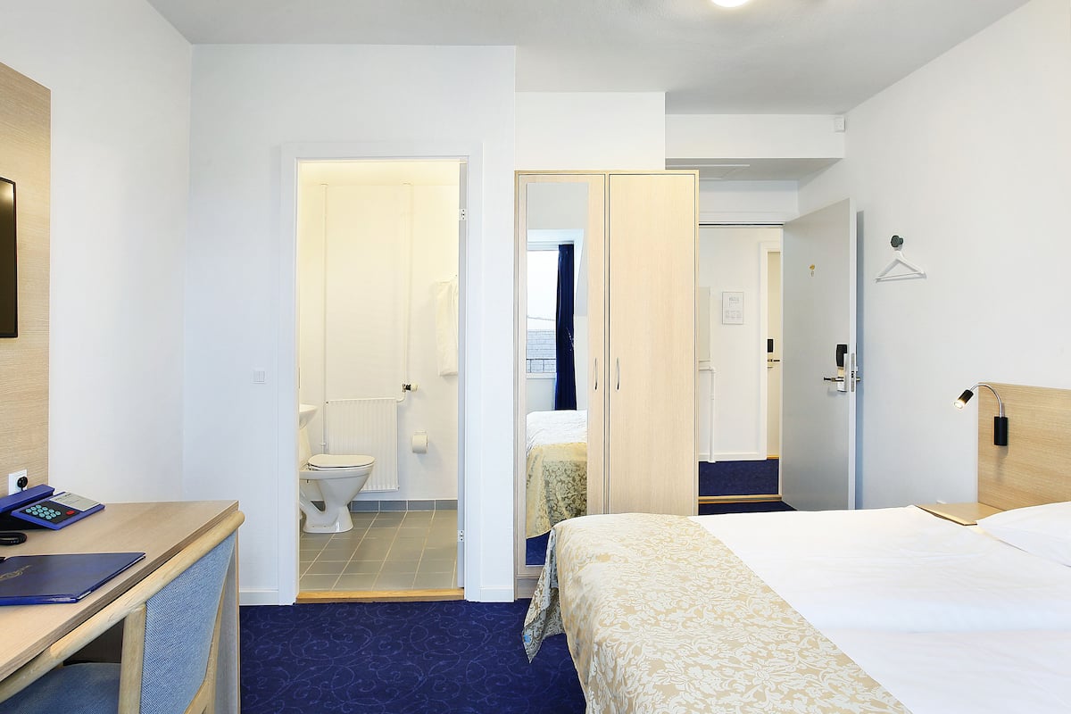 Hotellihuone, jossa on sänky, työpöytä, tuoli, vaatekaappi ja kylpyhuone, joka näkyy avoimesta ovesta.