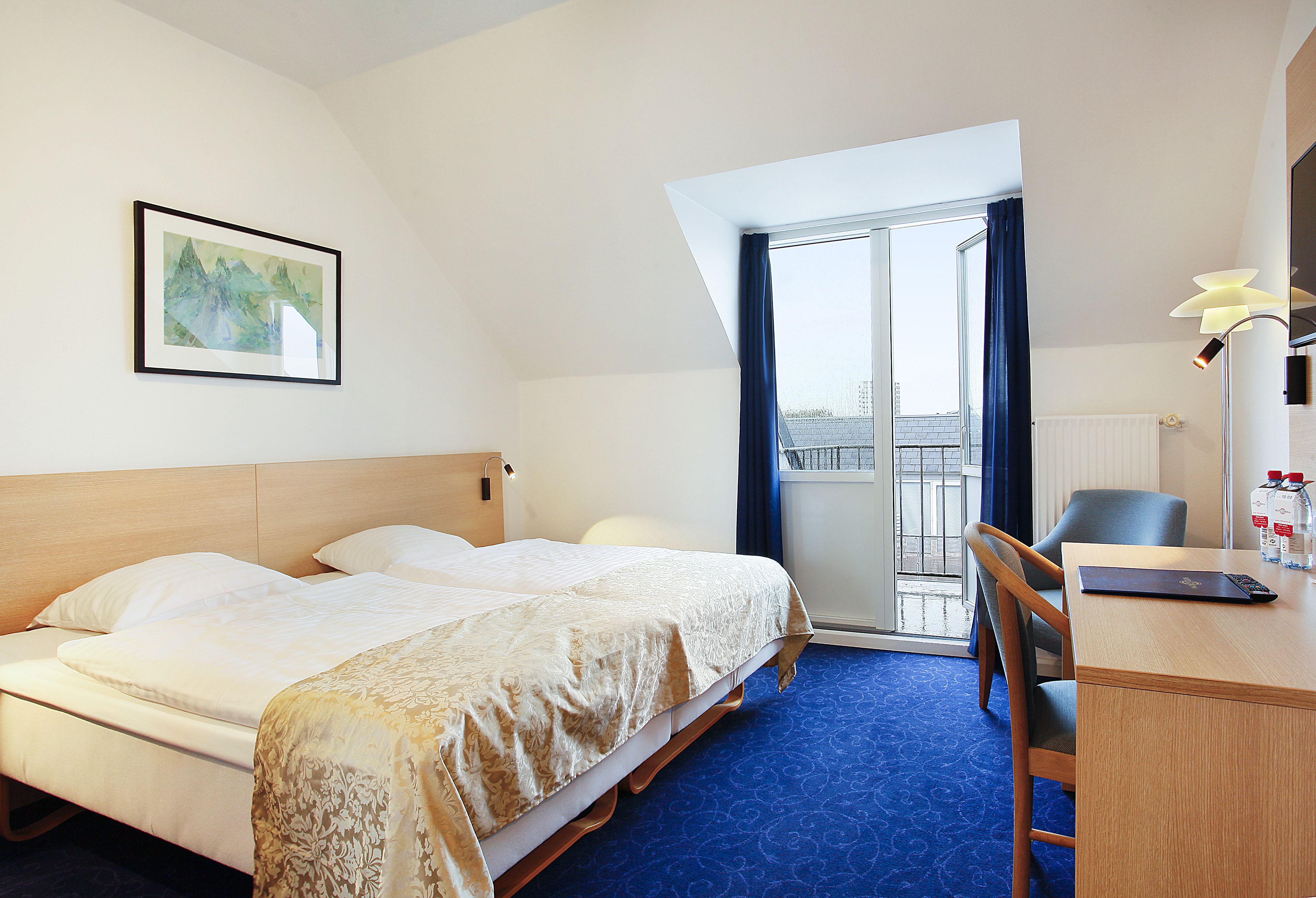 A hotel room with a bed, a desk, a chair, a window with curtains, and a framed picture on the wall.