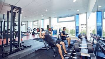 Un gimnasio con equipo de levantamiento de pesas, ventanas amplias y vistas a edificios.