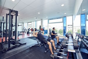 Un gimnasio con equipo de levantamiento de pesas, ventanas amplias y vistas a edificios.