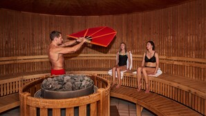 Eine Sauna mit Holzbänken, einer zentralen Feuerstelle und zwei Personen, die auf den Bänken sitzen.