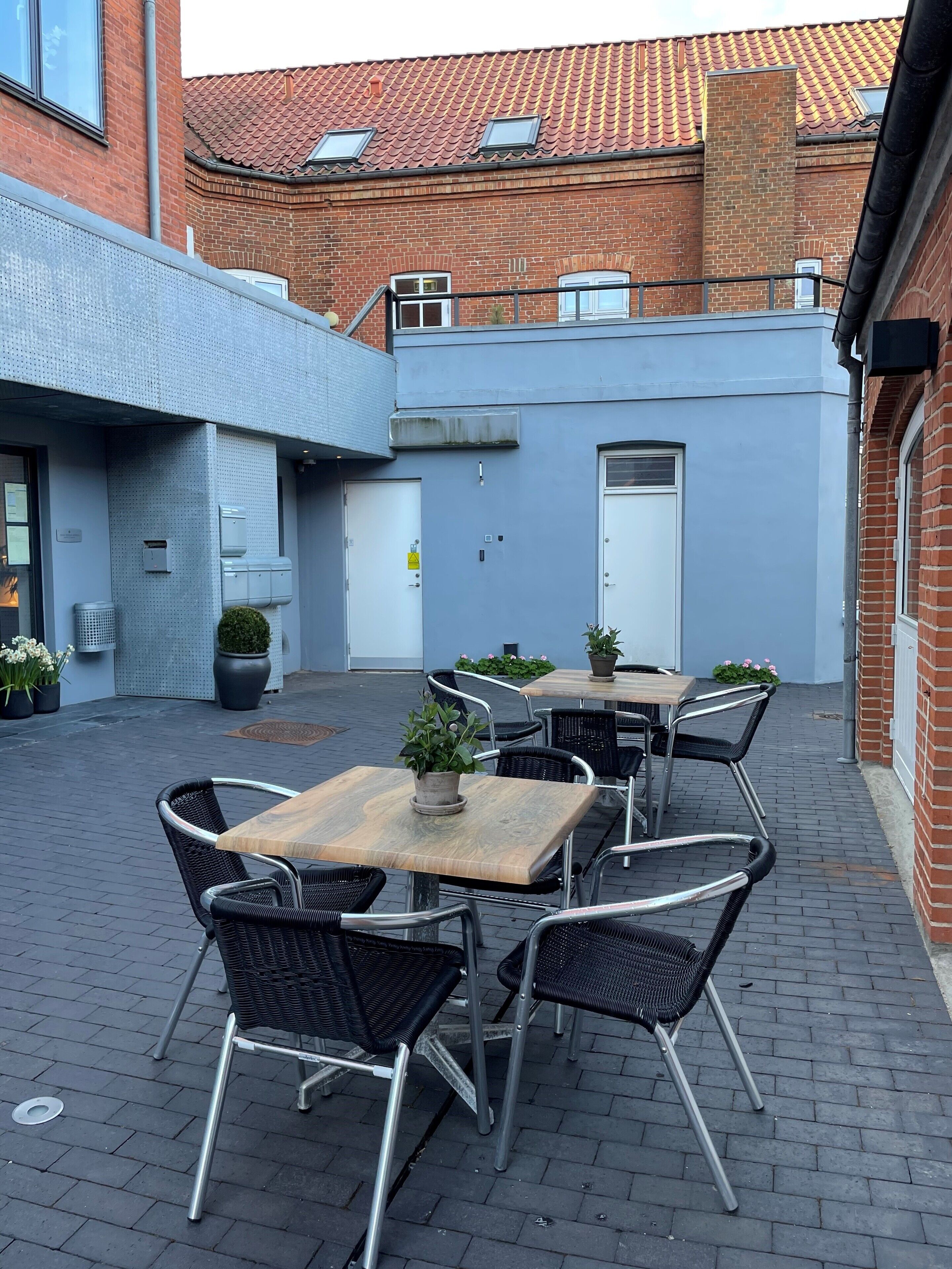 An outdoor patio area with a table and chairs, a potted plant, and a brick building in the background.