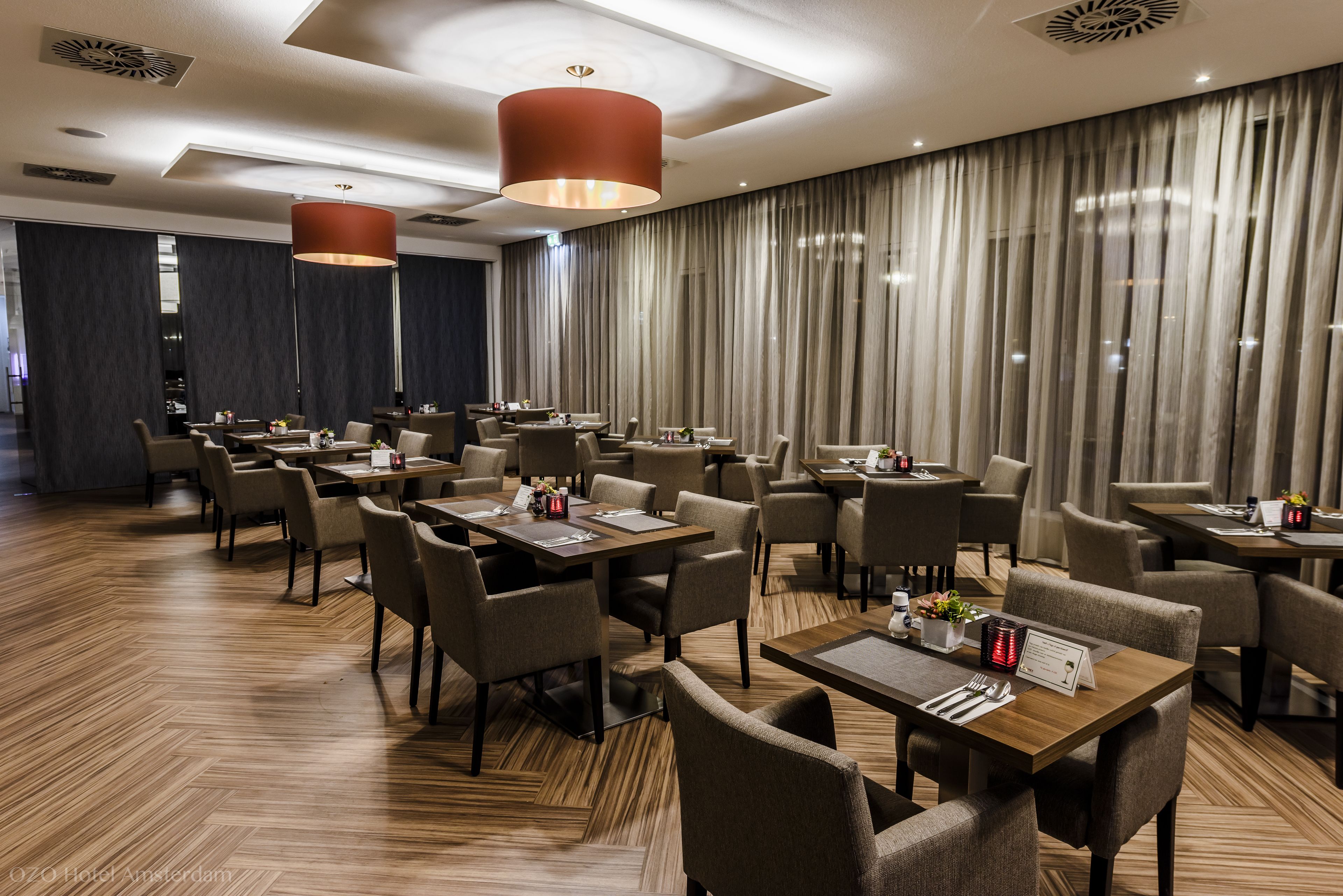 A modern hotel dining area with multiple tables and chairs, wooden flooring, and large windows with curtains.