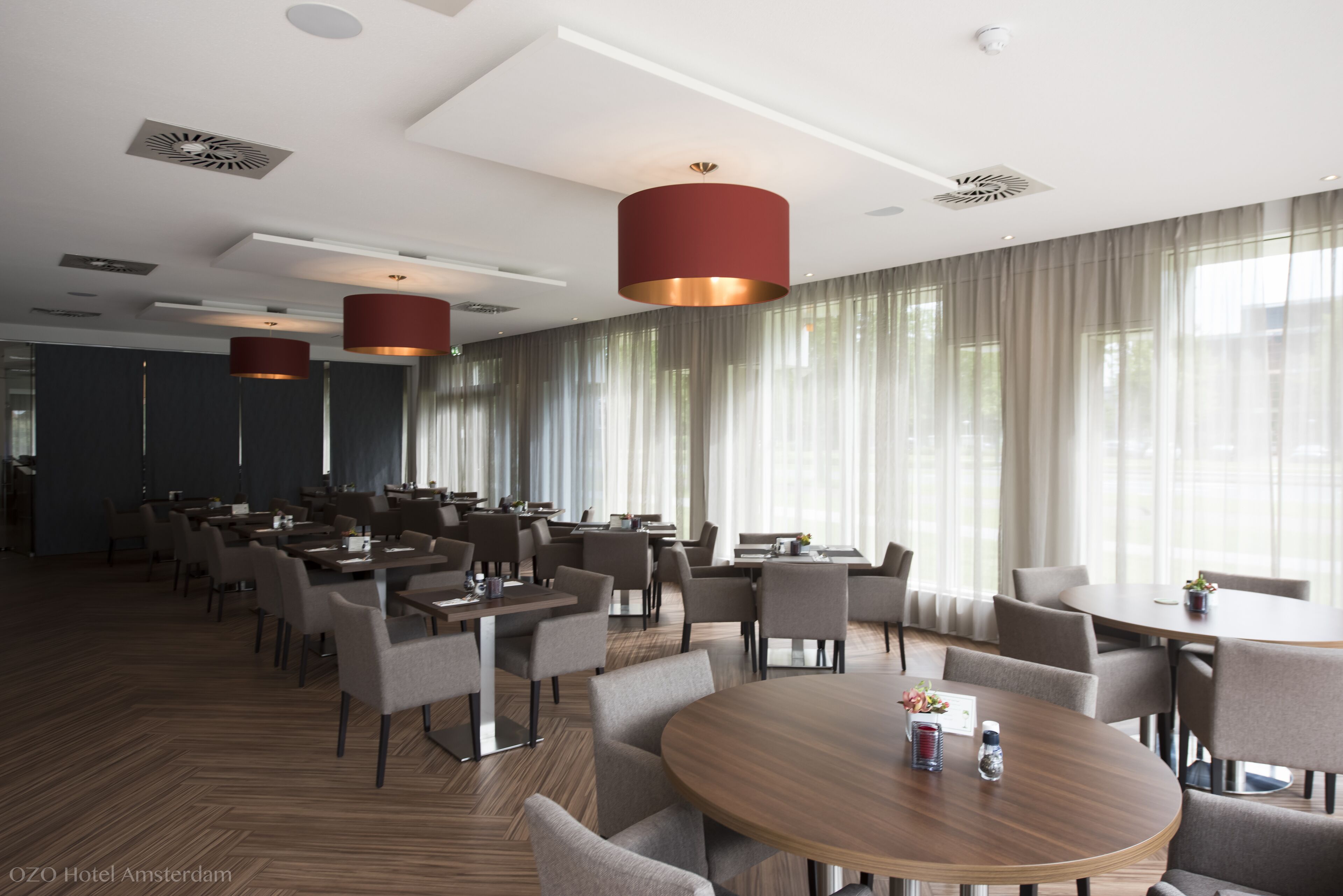 Un espace de restauration d’hôtel comprenant plusieurs tables et chaises, de grandes fenêtres avec des rideaux et un éclairage moderne suspendu.