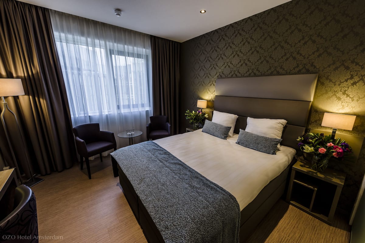 A hotel room with a large bed, two bedside tables with lamps, a chair, a small table, and a window with curtains.