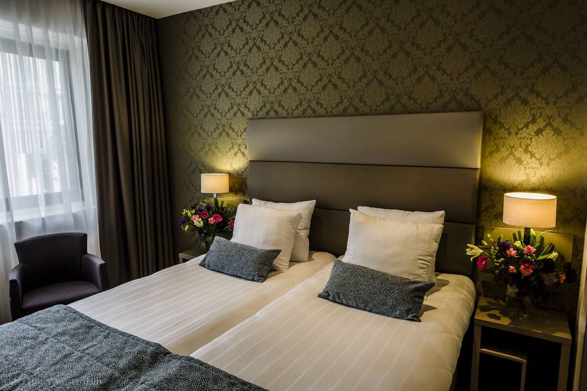A hotel room with two beds, a chair, and a nightstand with a lamp and flowers.