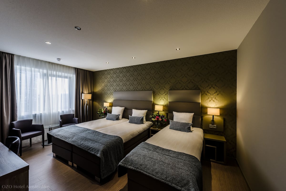 A hotel room with two beds, a desk, and a chair. There are lamps on the nightstands, and a window with curtains.