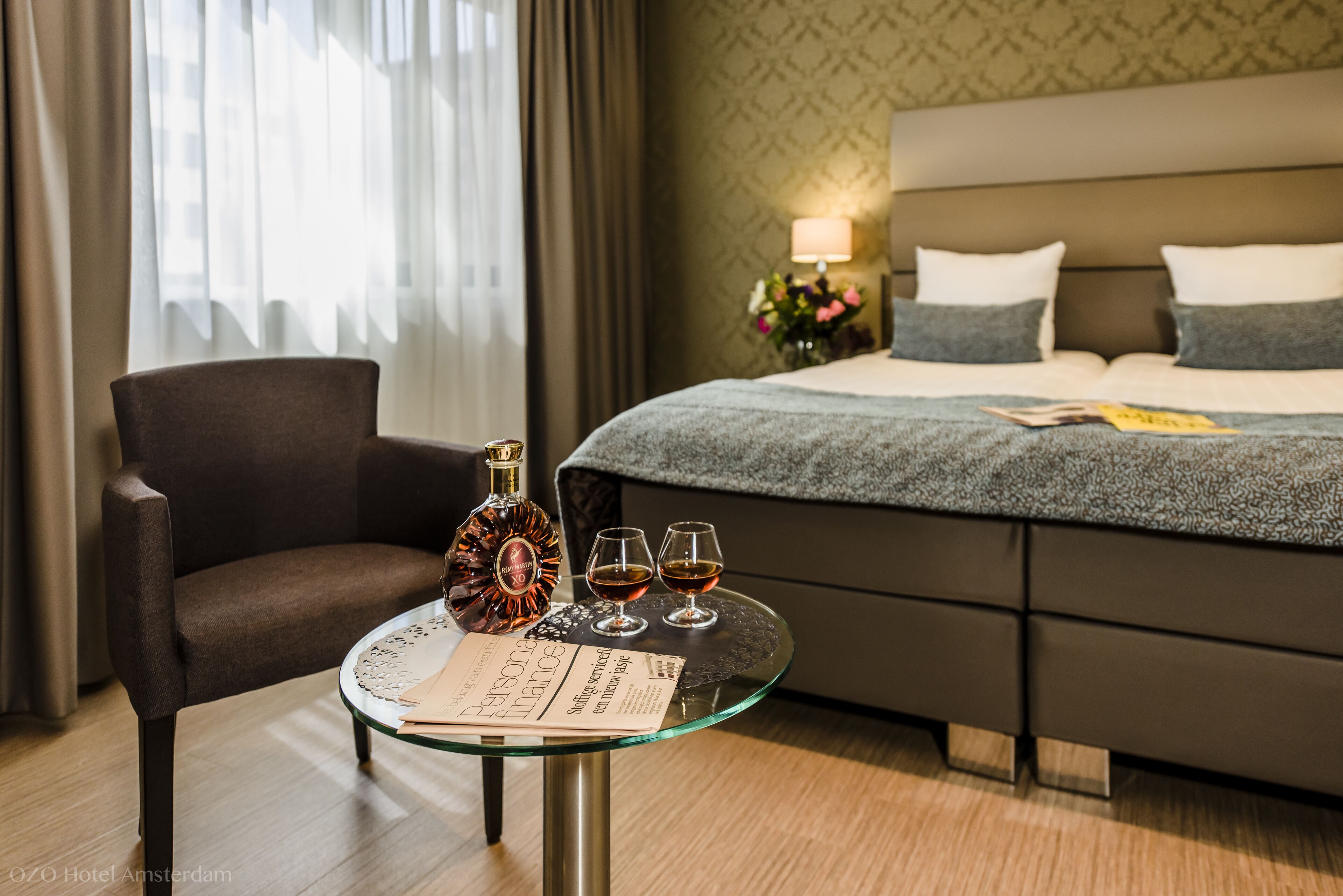 Habitación de hotel con cama, una mesa de vidrio, una botella y vasos, una silla y una lámpara.