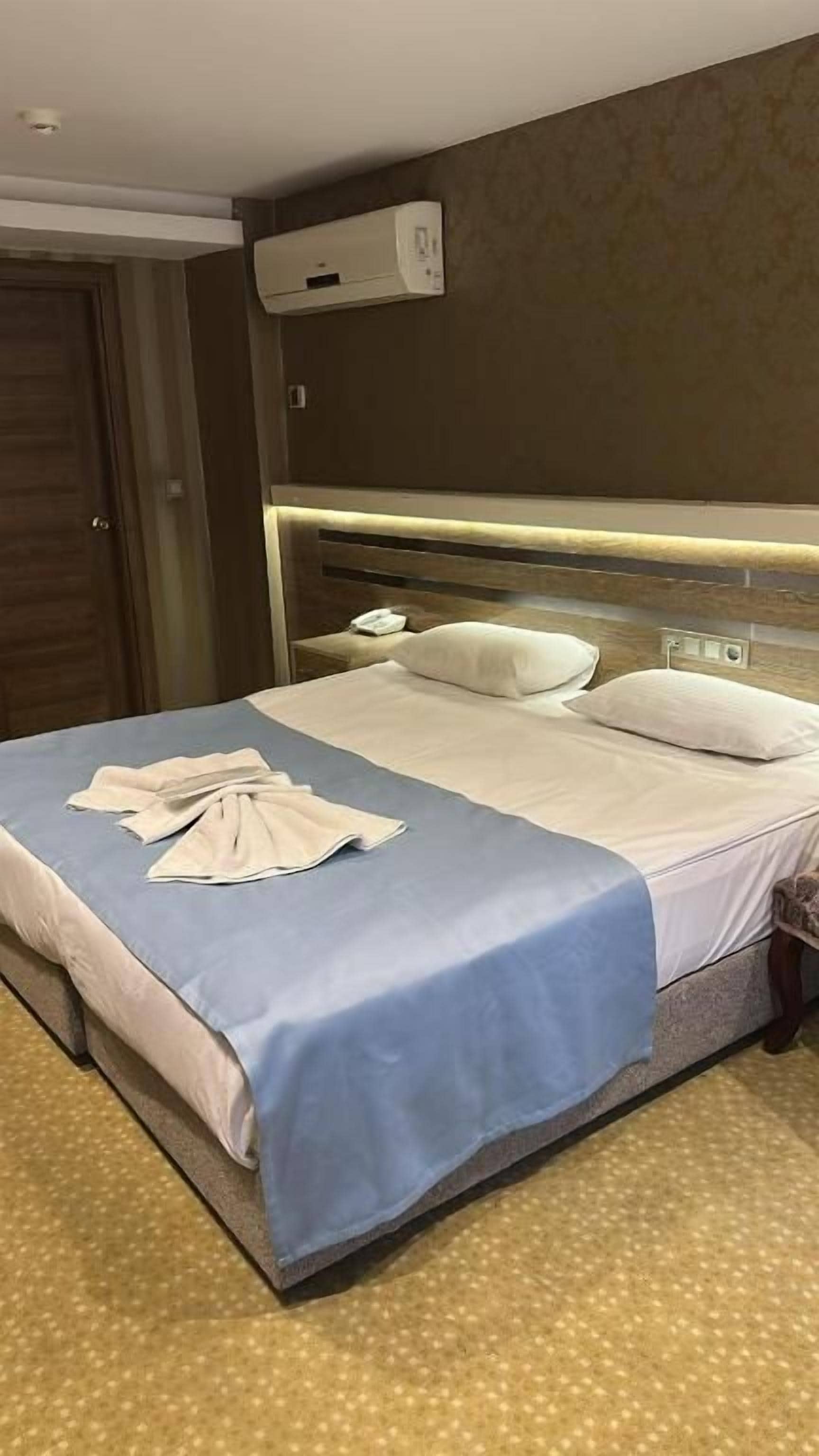 A hotel room with a bed, a bedside table, a wall-mounted air conditioner, and a patterned carpet.