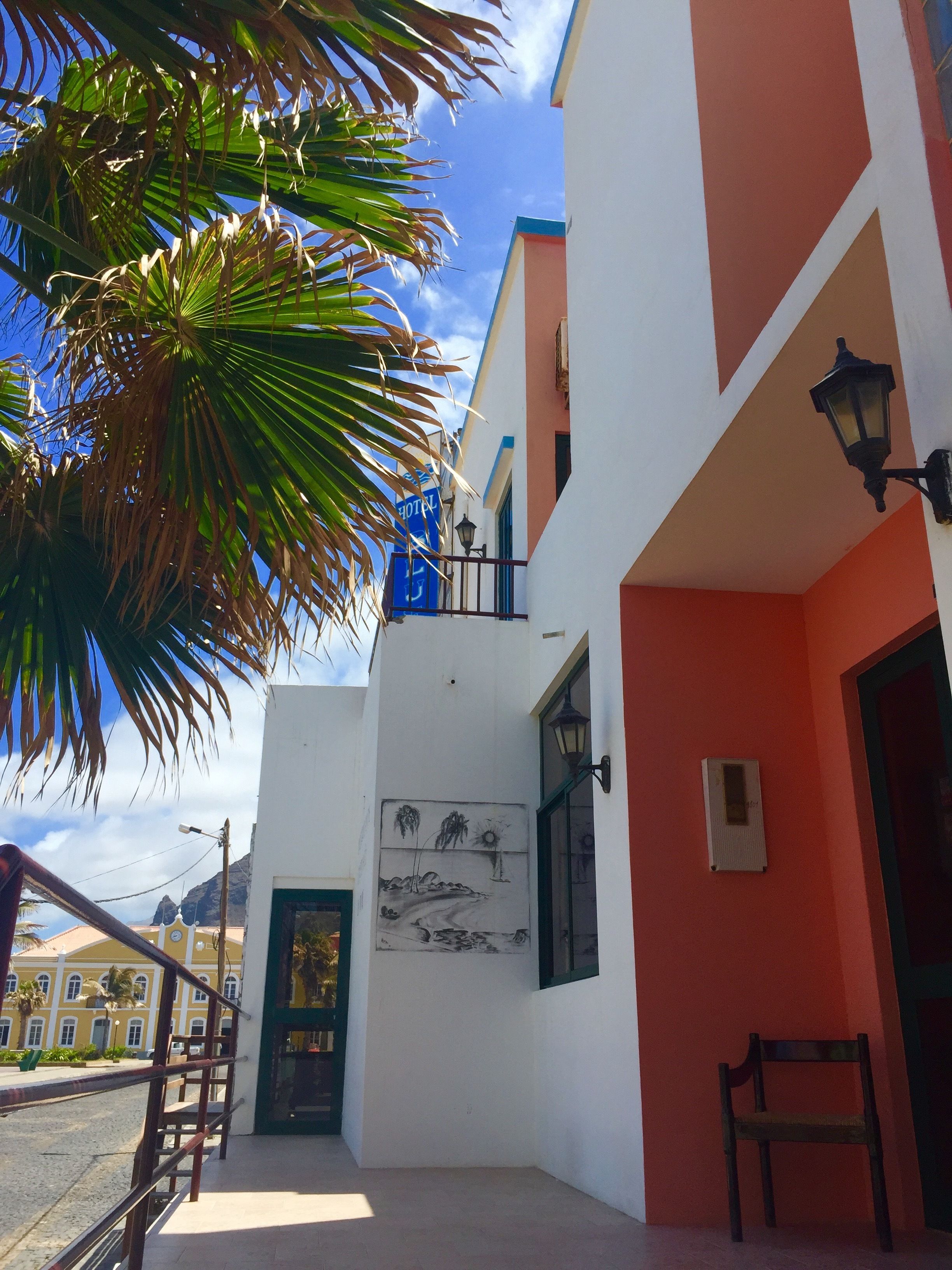 A hotel with a white and orange facade, a balcony with a metal railing, and a palm tree in the foreground.