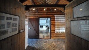Een houten interieur met een hoge plafond, foto’s in lijsten aan de muren en zicht op een moderne keuken door een open deur.