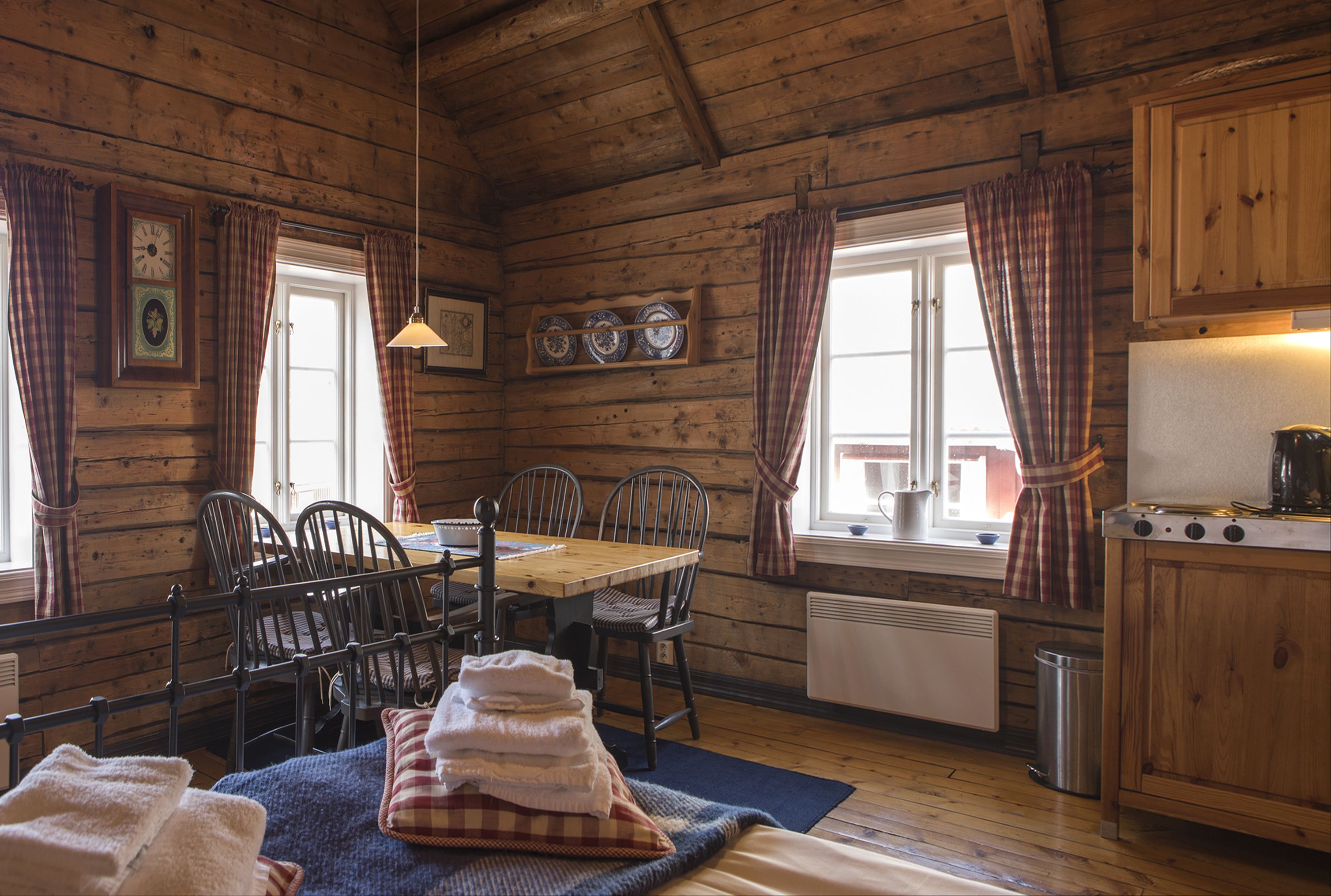 Une pièce confortable, aménagée comme un chalet, avec un intérieur en bois, une table à manger avec des chaises, un coin cuisine équipé d’une cuisinière et une fenêtre avec des rideaux.