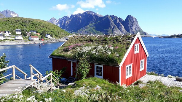 Een rood huis met een gazendak, een houten steiger en uitzicht op bergen en water.