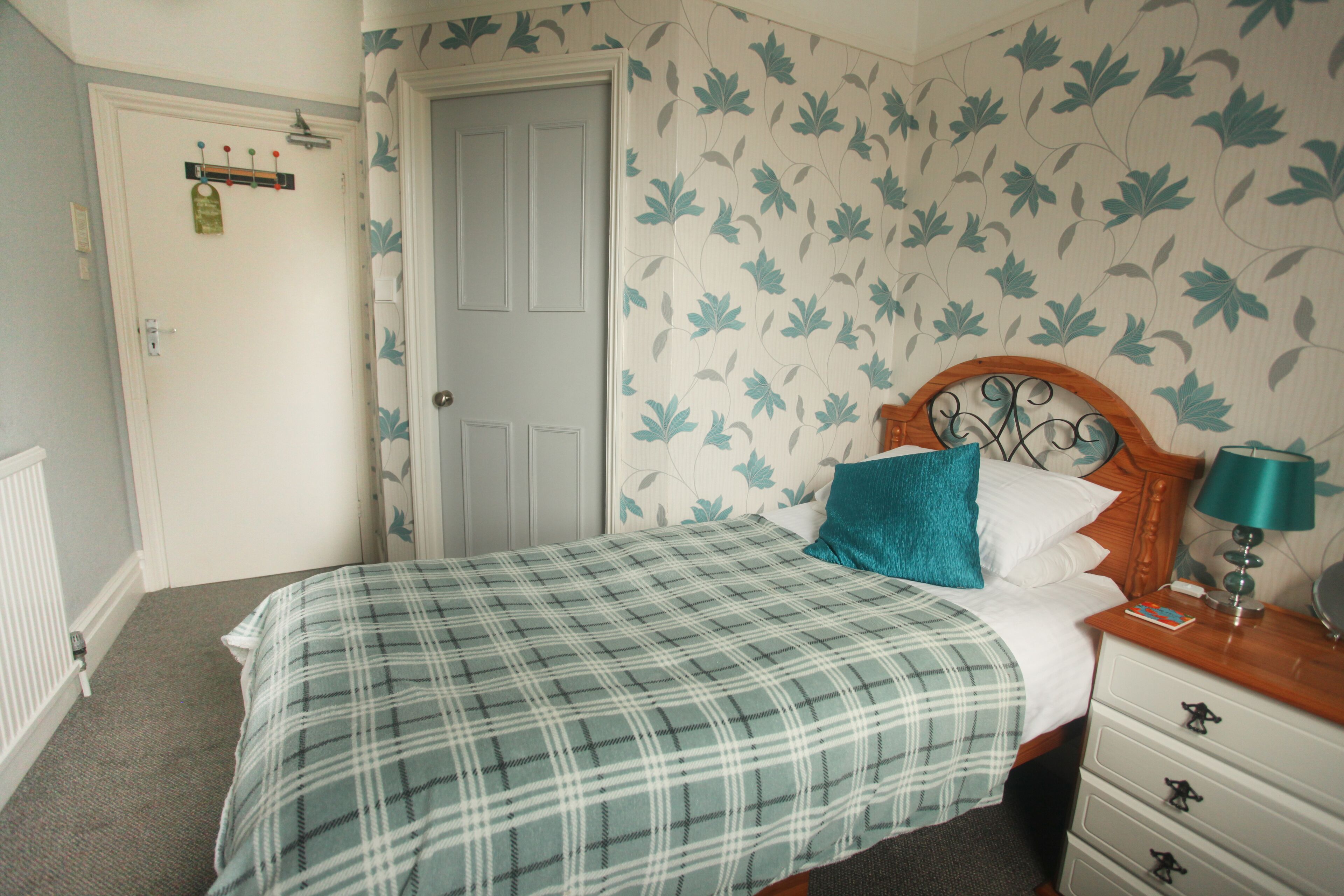 A bedroom with a plaid bedspread, a wooden headboard, a nightstand with a lamp, and a door with a coat rack.