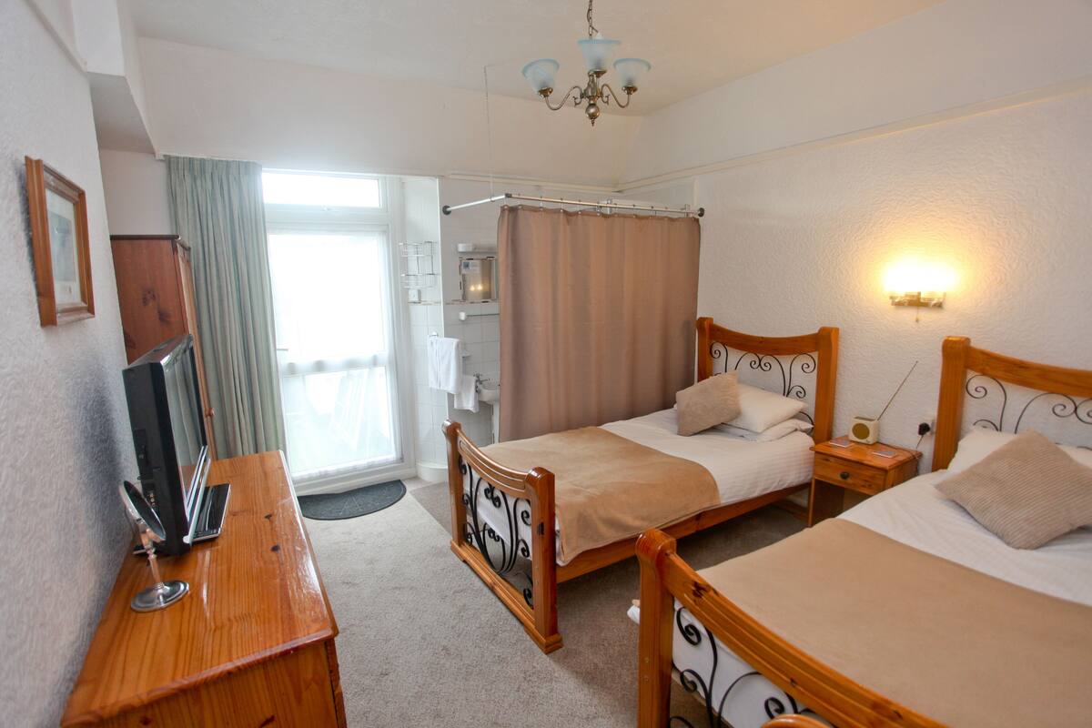 Habitación de hotel con dos camas individuales, un aparador de madera, un escritorio con computadora, televisor y una ventana con cortinas.