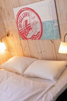 Una cama con ropa de cama blanca, una lámpara de pared y un cartel de un barco en la pared.