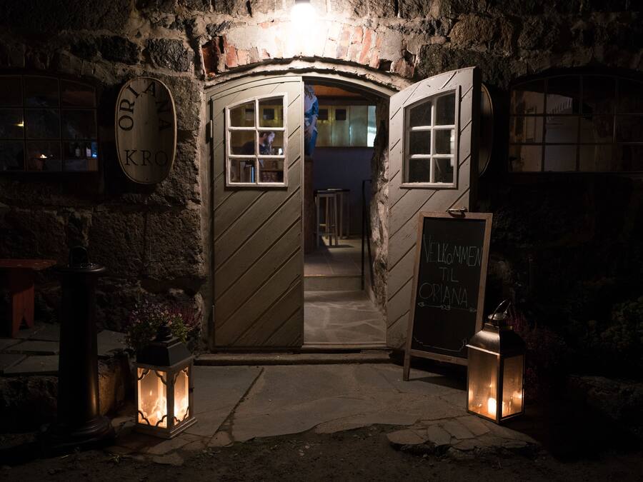 Entrada de un edificio de piedra con una puerta de madera, un letrero que dice 'Welcome to KRO' y un farol en el suelo.