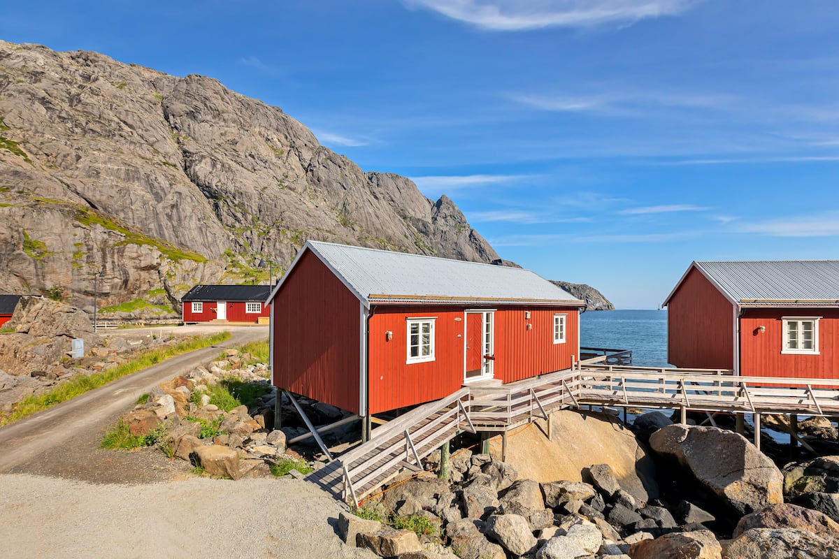 Two red wooden buildings with a metal roof, connected by a wooden walkway over rocks, near a rocky coastline.