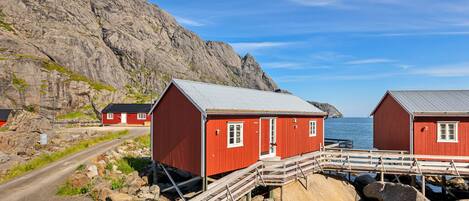 Two red wooden buildings with a metal roof, connected by a wooden walkway over rocks, near a rocky coastline.
