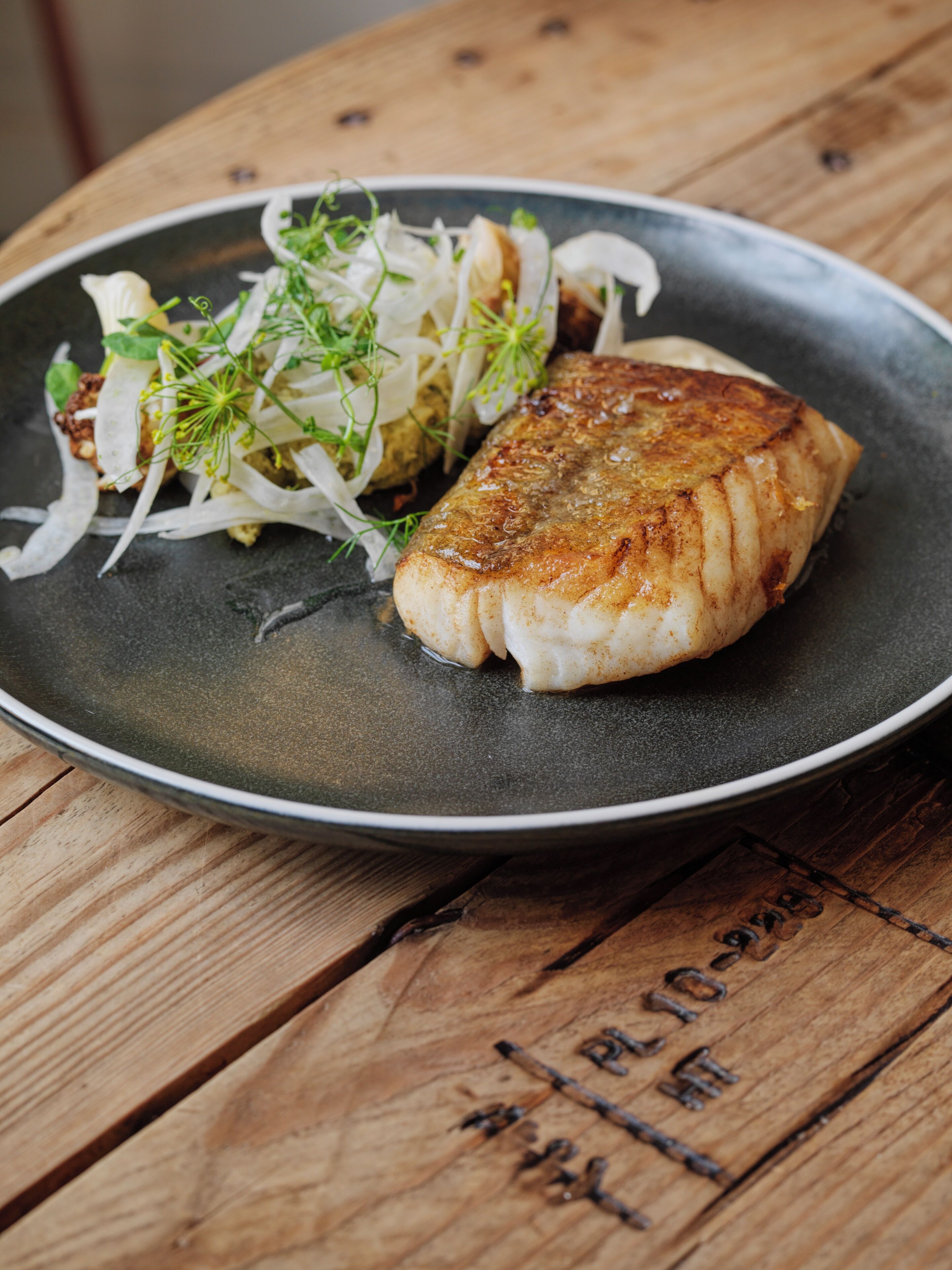 A plate of food with a piece of fish on a dark plate, accompanied by a side of greens and onions.