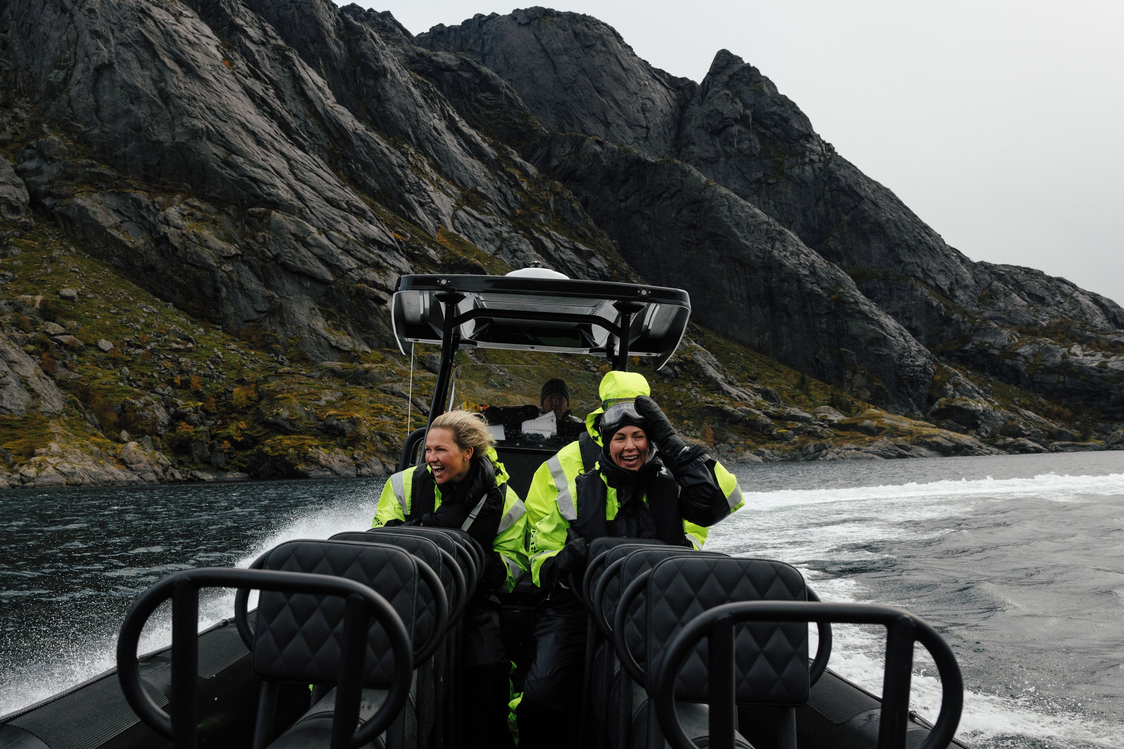 A boat with two people in safety gear, rocky mountains in the background, and a body of water.