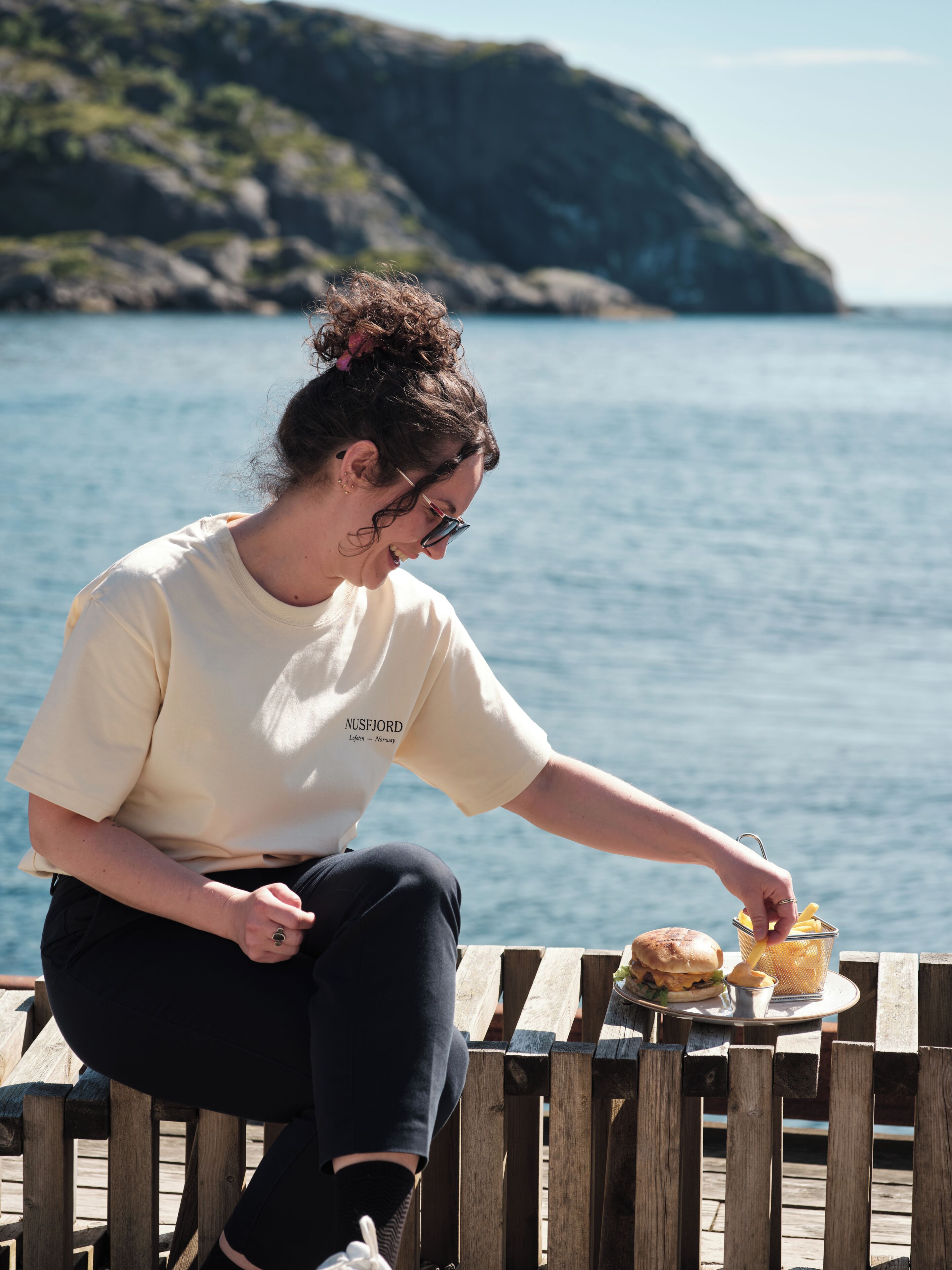Eine Person sitzt an einer hölzernen Bank am Wasser und isst ein Sandwich mit Pommes.