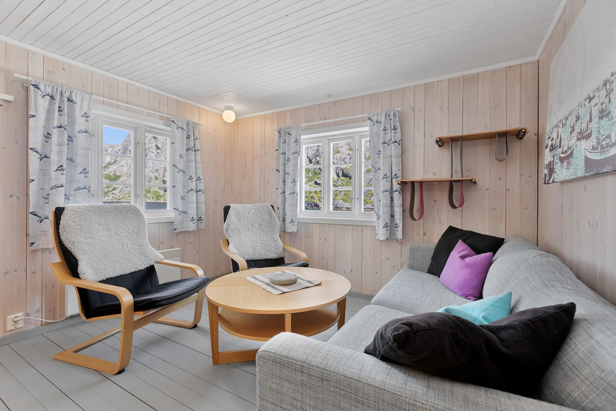 A cozy room with wooden walls, a round wooden table, two chairs, a sofa with cushions, and a window with curtains.