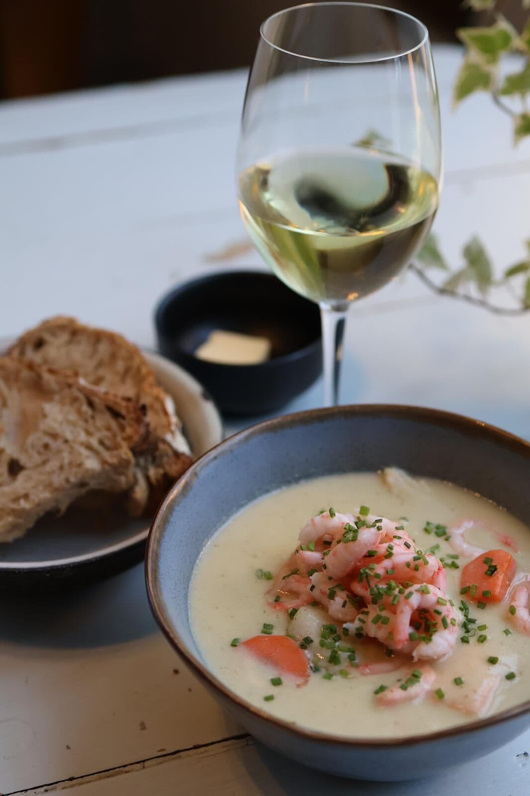 A bowl of seafood soup garnished with chives, a glass of white wine, and a slice of bread on a table.