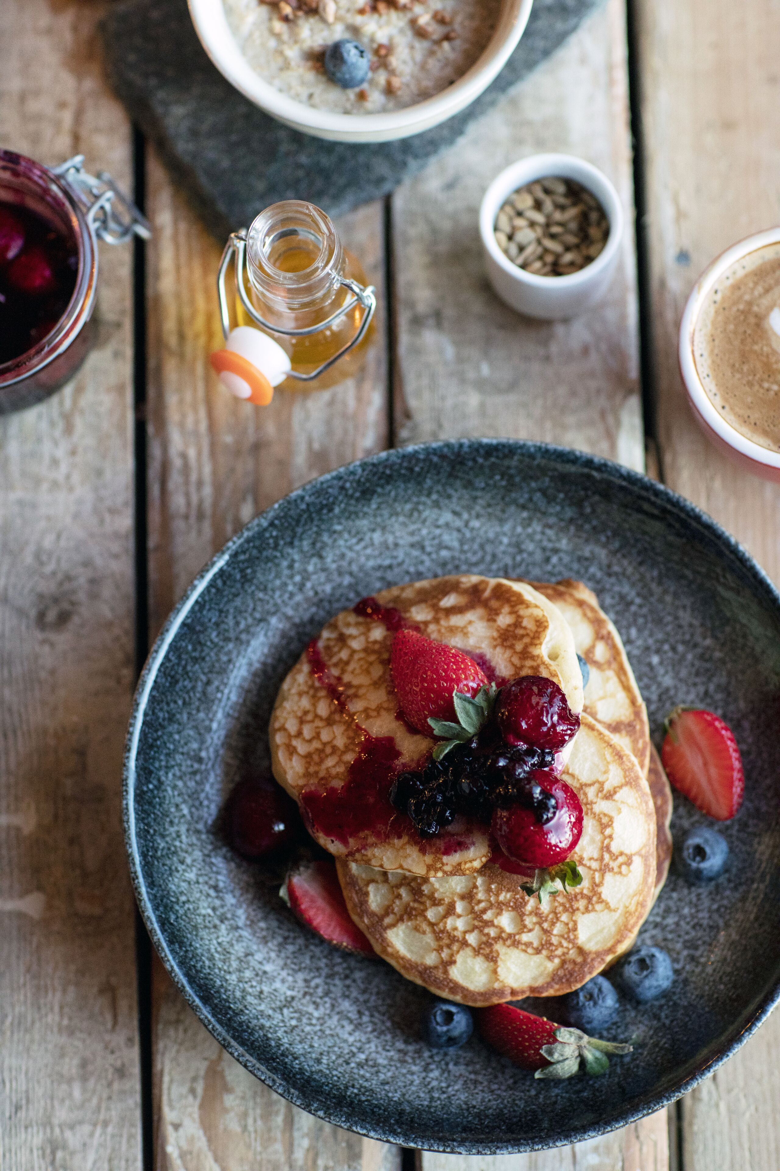 A breakfast setting with pancakes topped with berries and syrup, a small bowl of granola, and a bottle of honey on a rustic wooden table.