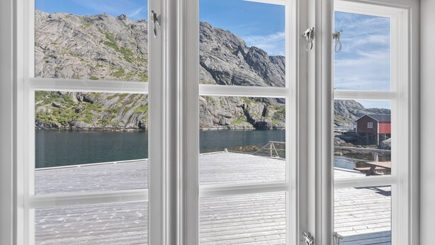 A view through a window of a wooden deck overlooking a rocky shoreline with a small building in the distance. - Lofoten Cottages (Flakstad)
