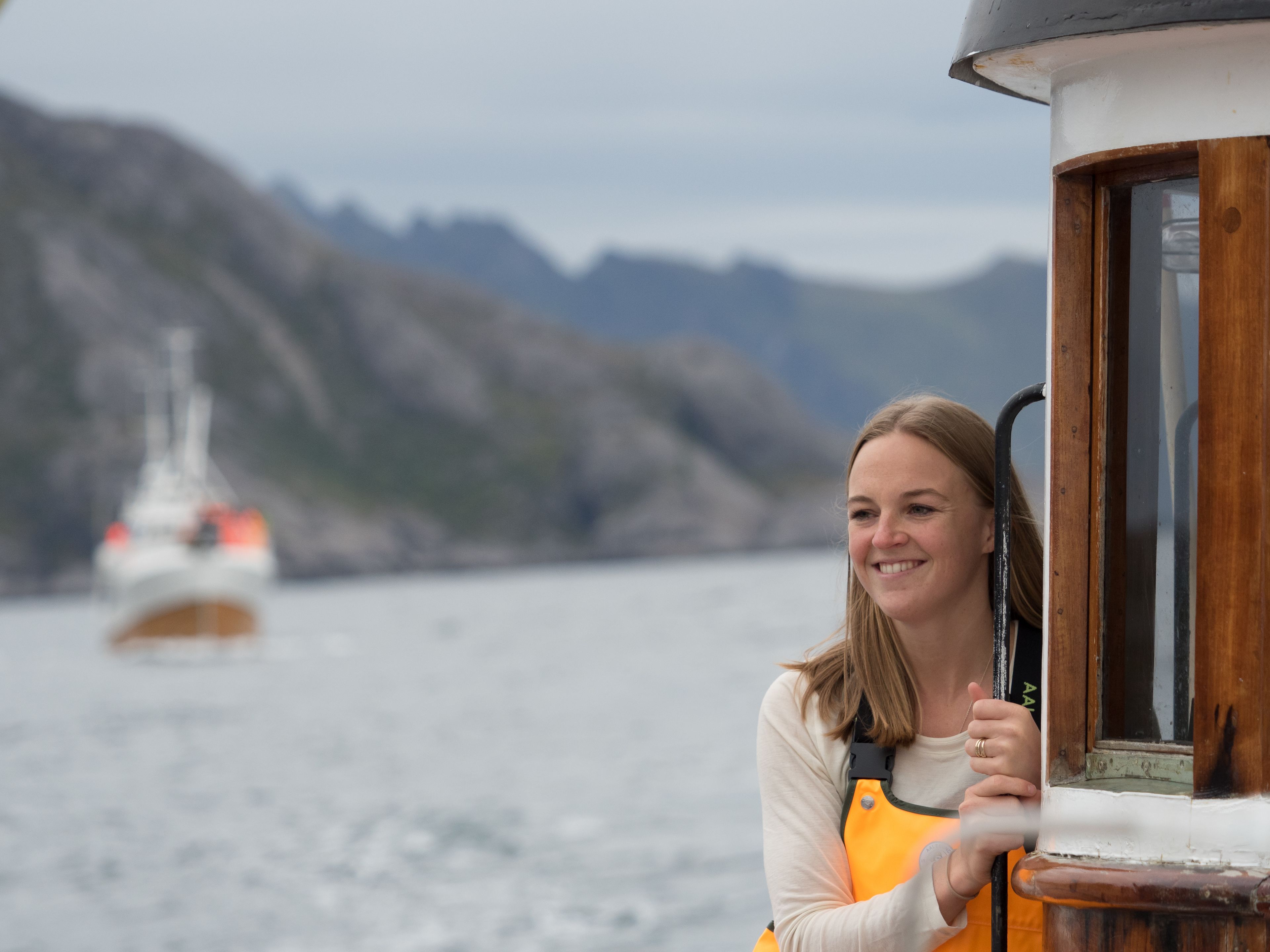 En person læner sig op ad et skibsråd, med et bjergrigt landskab og en båd i baggrunden.