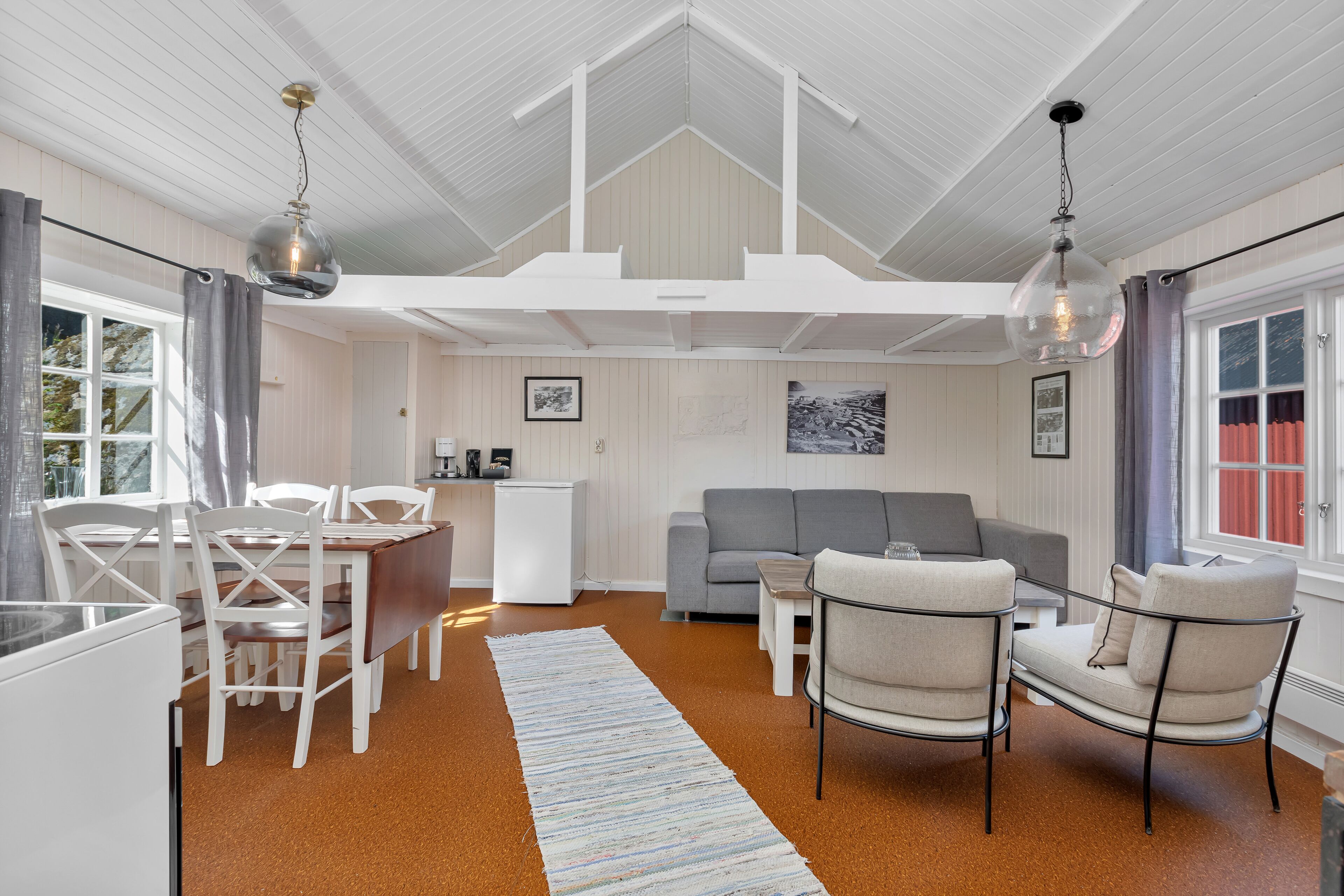Un vaste salon doté d’un haut plafond, d’un canapé gris, d’une table à manger avec des chaises, et d’un coin cuisine à l’arrière-plan.