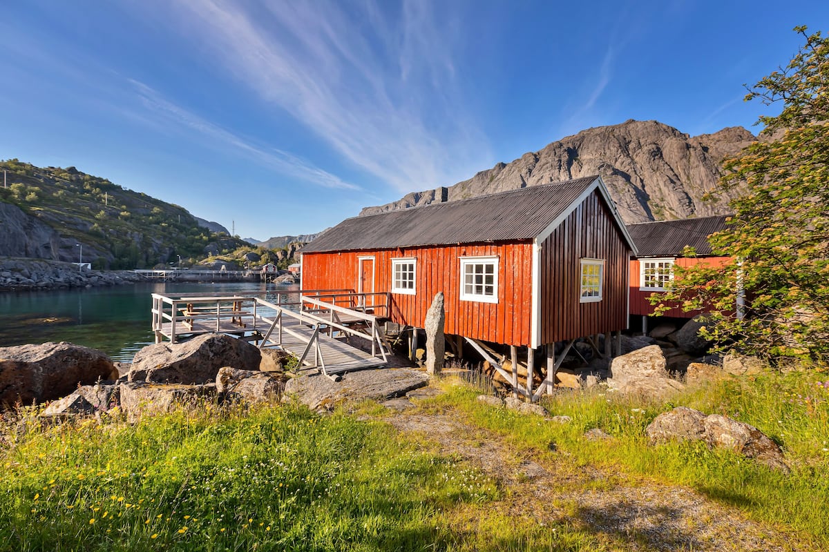 A red wooden house with a dock extending into a calm body of water, surrounded by rocky terrain and greenery.
