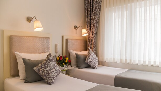 Une chambre d’hôtel avec deux lits, chacun doté d’une tête de lit, d’une table de chevet et d’une lampe. Il y a une fenêtre avec des rideaux légers et une vue sur l’extérieur.