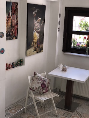 Een kamer met een witte tafel, een stoel met een bloemenkussen, een raam met uitzicht op planten en twee schilderijen aan de muur.