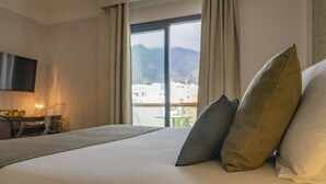 Habitación de hotel con cama, almohadas, escritorio con silla, televisor de pantalla plana y vistas a edificios y montañas a través de la ventana.