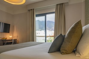 Habitación de hotel con cama, almohadas, escritorio con silla, televisor de pantalla plana y vistas a edificios y montañas a través de la ventana.