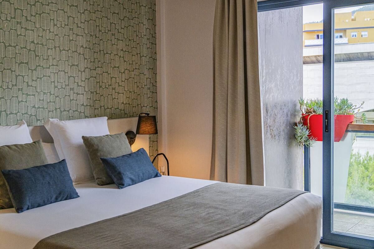 Una habitación de hotel moderna con una cama grande, una lámpara colgante, vista a través de un ventanal y una pared con diseño.