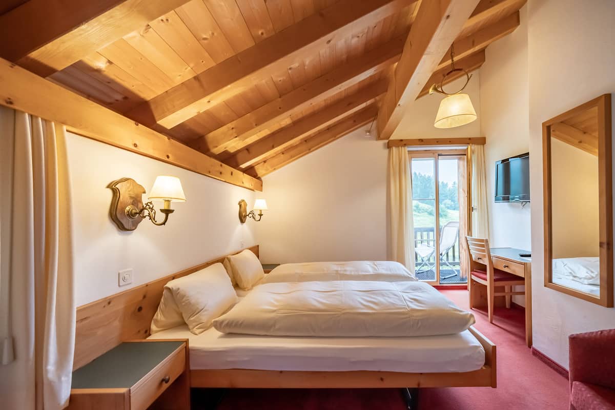 A bedroom with a wooden ceiling, a bed with white bedding, a desk with a chair, and a TV on the wall.