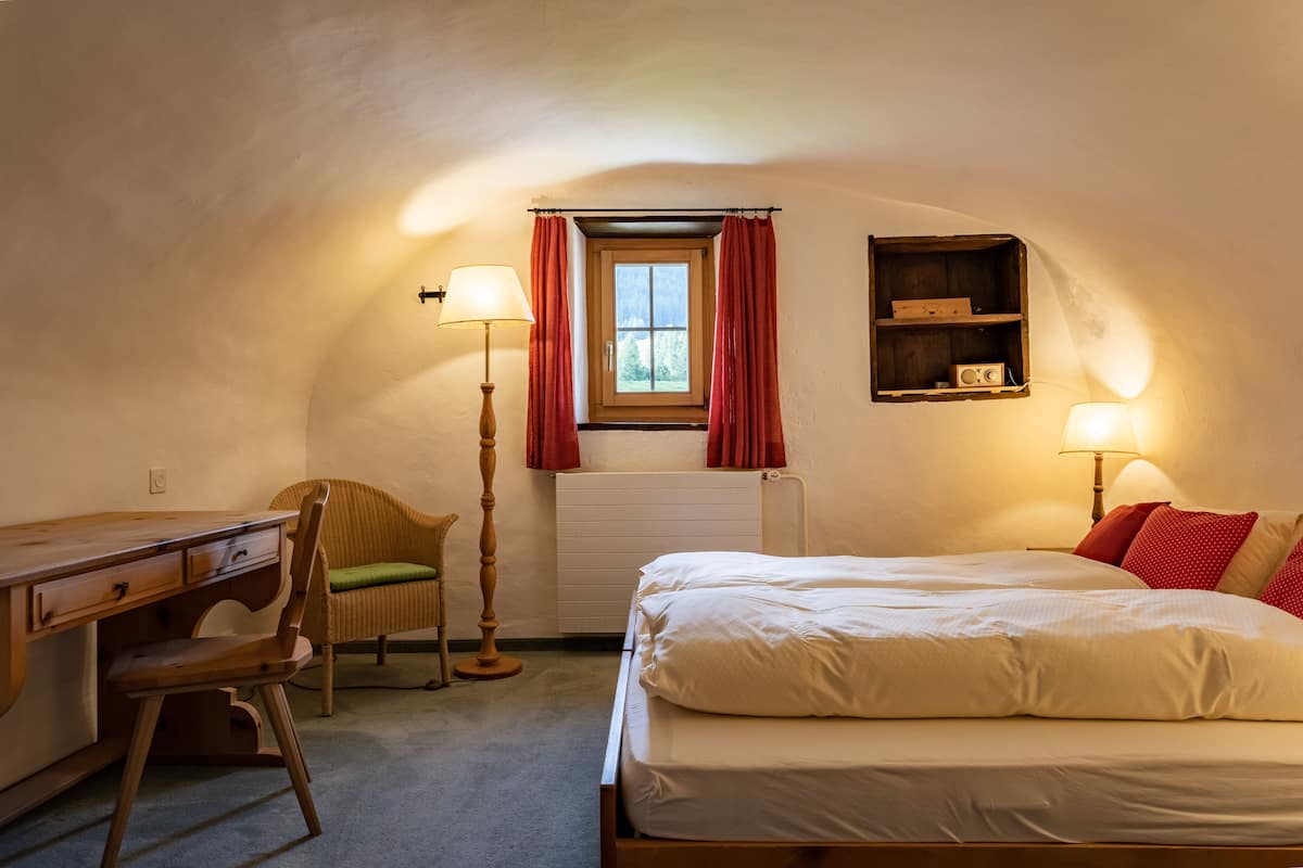 A bedroom with a bed, a desk, a chair, a lamp, a window with curtains, and a shelf with a radio.