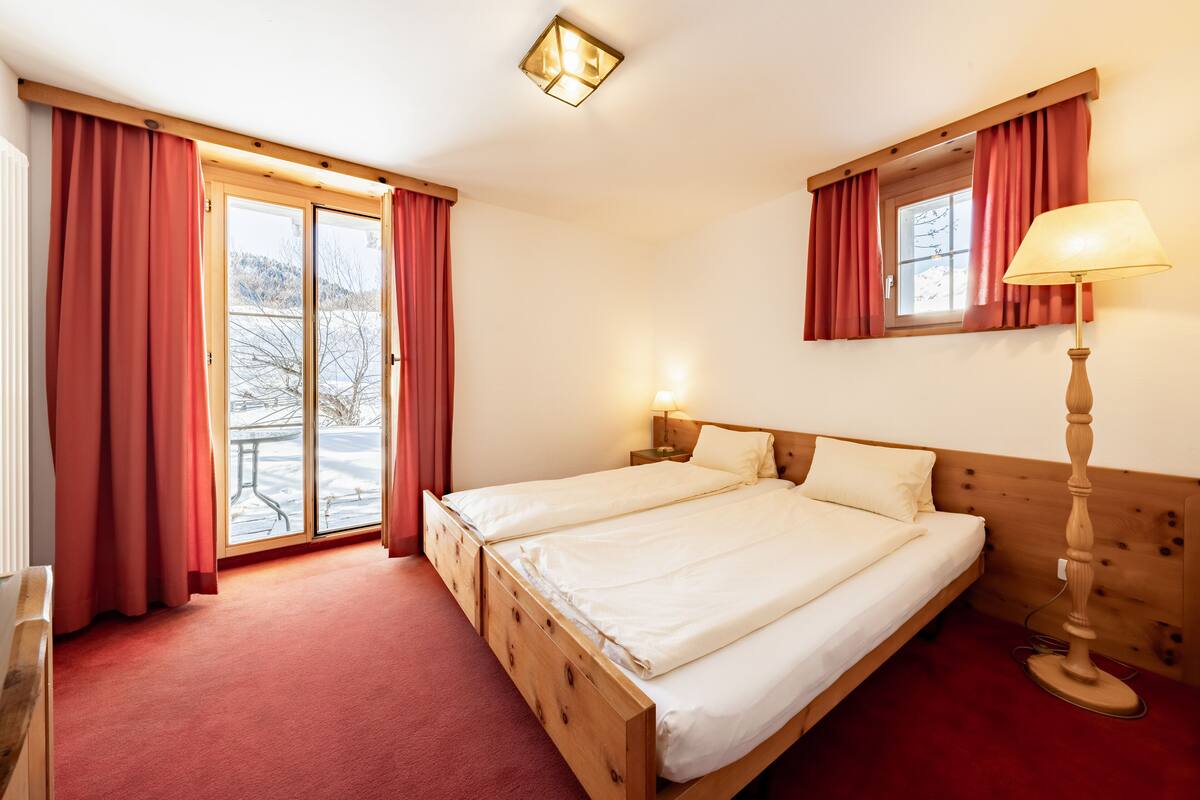 A double bed with a wooden headboard, a bedside lamp, and a window with red curtains.