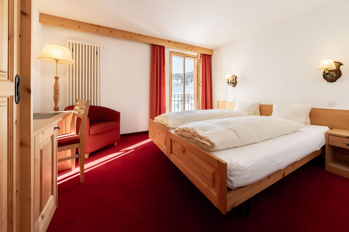 A hotel room with a wooden bed, a red chair, a wooden desk, a lamp, and a window with curtains.
