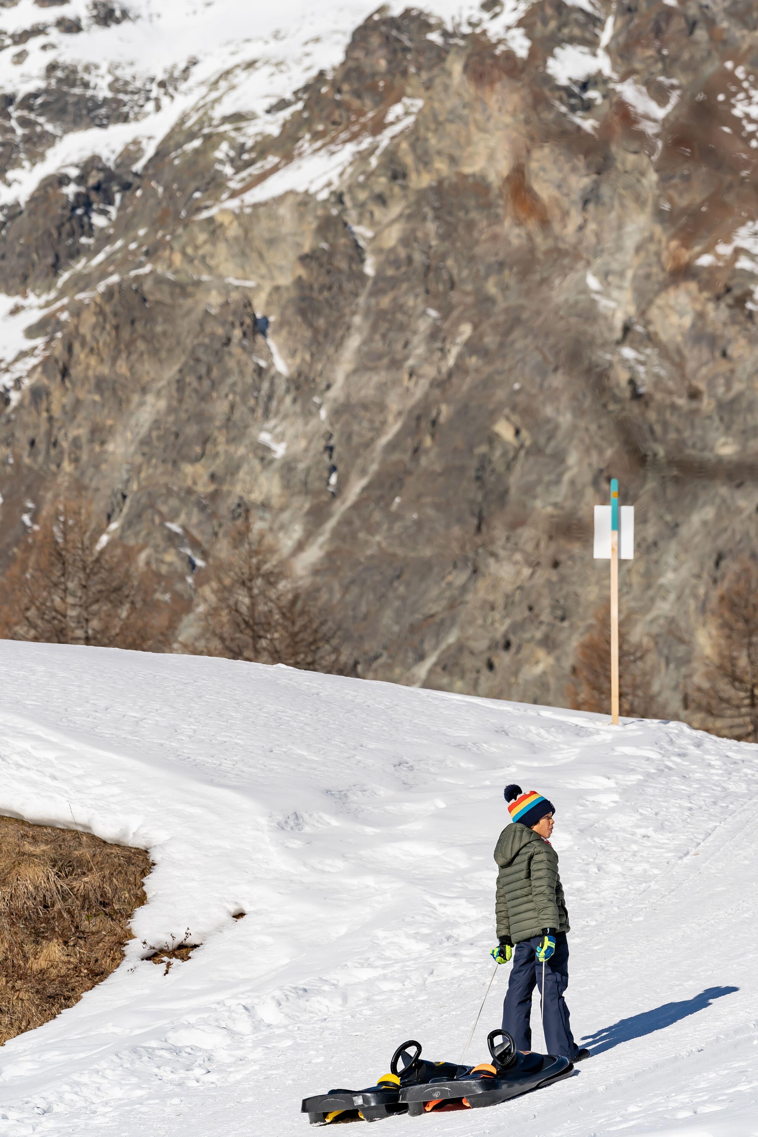 Eine Person steht mit einem Snowboard auf einem verschneiten Hang, trägt eine grüne Jacke und eine bunte Mütze.