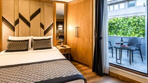 Habitación de hotel con una cama, un armario de madera, una mesita con un jarrón y un control remoto, y un balcón con sillas y una mesa.