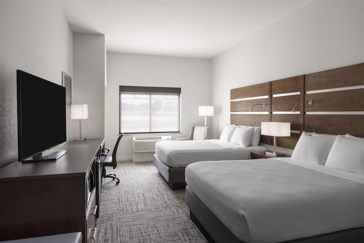 A hotel room with a TV, two beds, a desk, a chair, a window, and a wall-mounted light fixture.